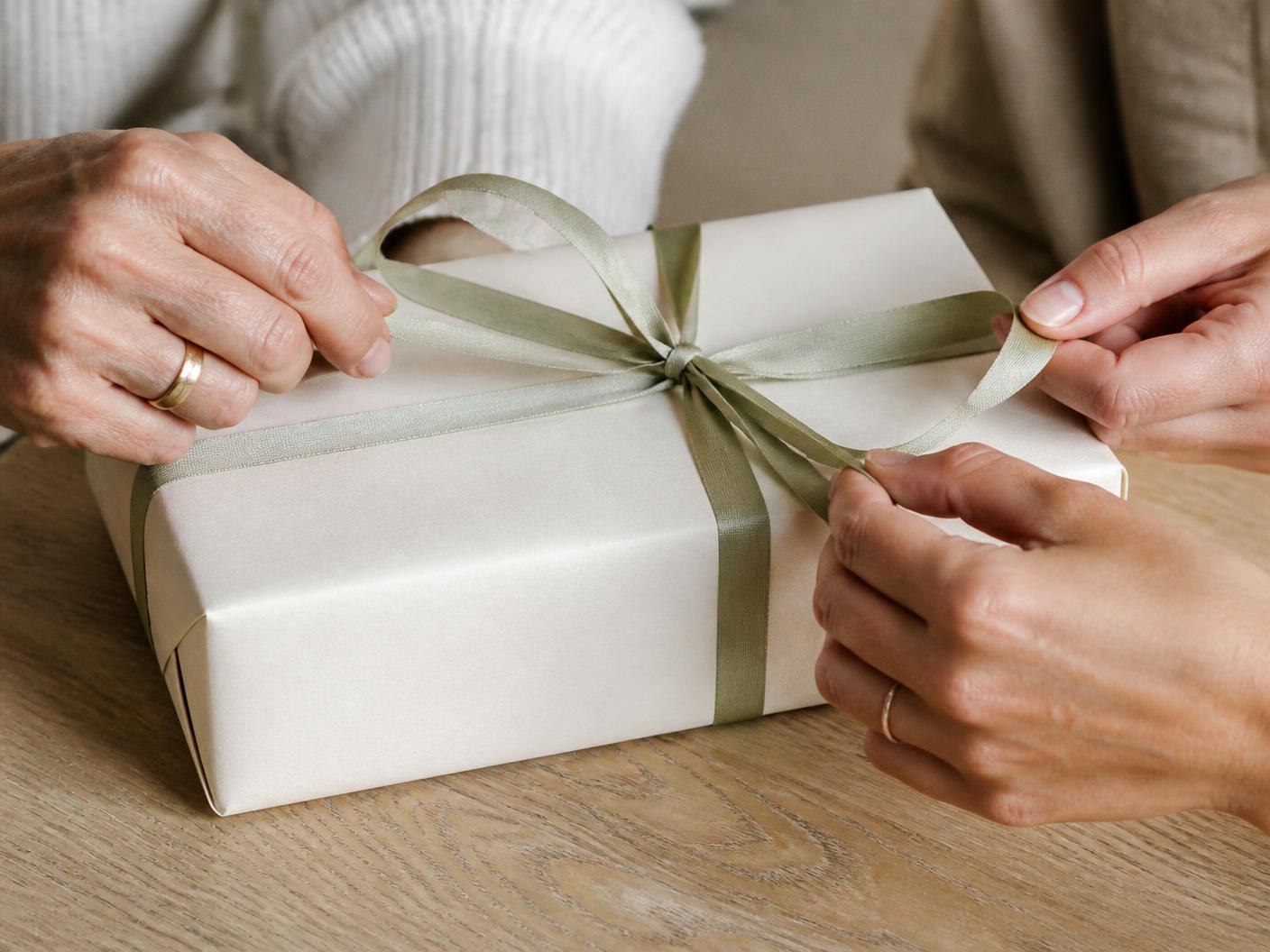 Two pairs of hands untying the sage ribbon on an ivory wrapped gift